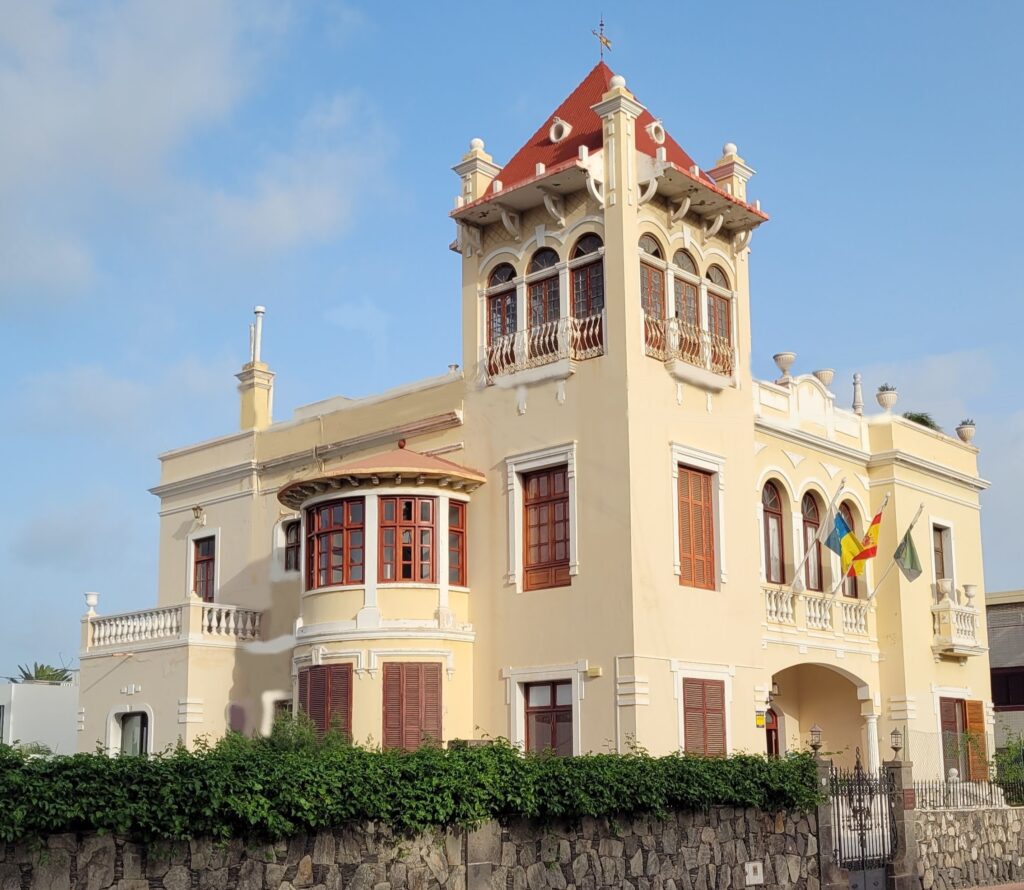 Casa de estilo arquitectónico colonial con detalles ornamentales, ubicada en un entorno urbano, rodeada de vegetación y con banderas ondeando en la fachada.